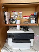 HP OfficeJet Pro 7740 printer with box packages of HP ink cartridges, Canon photo paper and other office supplies inside wooden cabinet shelf.