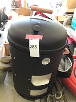 Front view of the black cylindrical MECO woodfired meat smoker with domed lid and wooden handle, thermometer on front, and vents with mesh coverings.