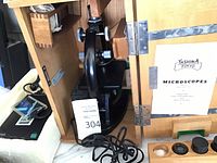 Black electric microscope inside wooden carrying case with metal hinges, showing microscope body, adjustment knobs, and power cord