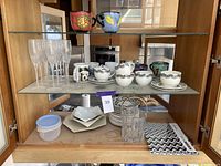 Cabinet shelf displaying crystal champagne glasses, Missoni cappuccino mugs and saucers, white geometric plates and bowl underneath, black and white patterned napkins.