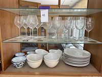 Glass shelving with Royal Doulton white dinner plates and bowls, blue and white Asian rice bowls, and several wine glasses on top shelf, including Riedel and Lakers glasses.