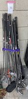 Photo of a bundle of assorted golf clubs with various heads and black grips tied with a purple strap leaned against a wall.