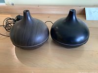 Photo of two aromatherapy diffusers, one solid black and one wood grain finish, placed side by side on a wooden surface with cords visible behind.
