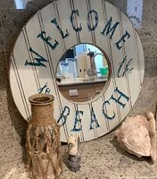 Photo showing round beach-themed wall mirror, jute-wrapped glass vase, wooden seagull figure, and seashell arranged together