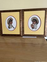 Two framed cross stitch artworks of Mexican children side by side on a wooden surface.