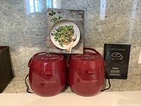 Two Curtis Stone red multi cookers placed side by side with their power cords visible. Behind them are two books: 'The Ultimate Multi Cooker Cookbook' and a Curtis Stone multi cooker manual booklet. The photo shows the full set included in the lot.