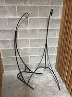 Two black metal garden flower basket stands with tripod bases and curved hooks for hanging baskets. Stands positioned indoors against a concrete block wall.
