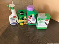 Four garden supply products on dark surface: large green container of Miracle-Gro Shake 'n Feed with purple lid, smaller Miracle-Gro Water Soluble Plant Food box, Safer's Slug & Snail bait container, and Safer's Critter Ridder spray bottle.