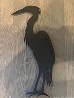 Solid black metal heron silhouette wall art piece photographed on wooden floor, showing whole body from beak to feet with detailed feather edges.