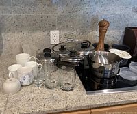 Wide shot of kitchen countertop showing various stainless steel pots with lids, clear glass tumblers, ceramic mugs, wooden pepper grinder, and kitchen accessories like egg poacher and lids.
