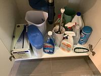 Image of a cupboard shelf with assorted cleaning and household items including sprays, wipes, brushes, tissues, and plastic containers.
