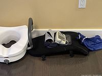 Photo showing a white plastic toilet riser with attached gray armrests, a digital blood pressure monitor with cuff and blue protective cover, and a black padded back support pad for chair.