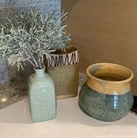 Photo of three pottery items: a green rectangular etched vase with plant, a beige square vase with brown drip glaze, and a round green and tan glazed pot.