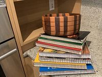 Photo of a kitchen counter shelf with seven spiral-bound Ridge cookbooks stacked horizontally and a large two-tone wooden turned bowl placed on top.