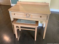 Wooden small desk with one large drawer and decorative metal ring pull, with matching wooden stool having a woven rattan seat below the desk.