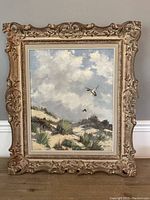 Oil painting in ornate frame leaning against wall showing full artwork of beach scene with birds.