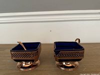 Pair of blue glass and brass creamer and sugar bowl seen from the front on a wooden table with a plain wall background.