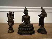 Three bronze statues arranged side by side: standing Ganesha at left, seated Buddha center, standing male deity at right