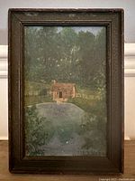 Full view of framed landscape painting showing a cabin by a pond set against a wooded background with a wooden frame.