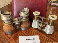 Two pairs of antique opera glasses with a red leather carrying case, showing one pair with mother-of-pearl inlay and the other pair covered in worn brown leather.