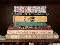 Stack of six cartography books on wooden shelf, titles and authors clearly visible.