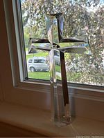 Front view of tall crystal cross standing on window sill with reflections of outdoor scenery visible through clear crystal.