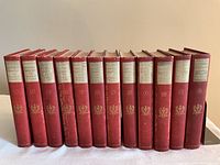 Front view of all 12 red hardcover volumes standing side by side, spines numbered I–XII with white labels and gold crest.