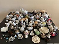 Wide view of the large assortment of miniature porcelain tea items arranged on a table showing various teapots, cups, saucers, and pitchers with floral and decorative patterns