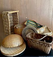 Wide view of seven various woven baskets showing different shapes, sizes, and weave styles arranged on a black surface against wood paneled wall.