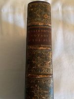 Spine of the book showing the embossed title 'McClintock’s Voyage of The Fox' on a red leather label with ornate gold floral designs above and below.