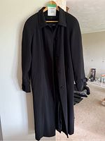 Full-length black trench coat hanging on a door, showing front and sleeve detail.