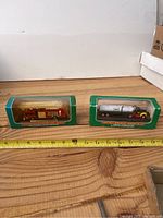 Two Hess miniature trucks displayed in original green and clear plastic boxes, placed side by side with a measuring tape in front showing approx 12 inches length total.