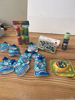 Overview photo displaying all items in the lot on wooden surface: nine small blue bubble containers, triple pack of slime tubes, game with spinner and colorful marbles, and cookie-shaped game controllers packaged.