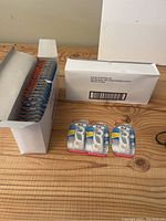 Two white boxes on a wooden surface, one open showing multiple blister packs of Tek dental floss picks, the other closed. Additional loose packs of floss picks are laid out.