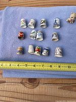 Photo showing 13 different decorative thimbles arranged on a blue cloth with a measuring tape beneath them for scale.
