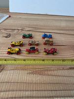 Eight miniature vintage die-cast cars arranged on a wooden surface with a measuring tape showing sizes around 2 inches.
