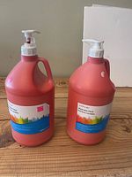 Two one-gallon plastic bottles of salmon pink washable paint with pump dispensers on a wooden surface.