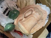 Box containing pinkish large handled pitcher, matching large charger plate, two small green ceramic frog figurines surrounded by white plastic bags.