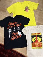 All three t-shirts laid out flat showing full graphic designs: black Panic! At The Disco, yellow Keith Haring, white Star Trek Mirror Mirror