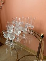 Set of 14 vintage glass goblets and flutes arranged on a glass table against a peach wall.