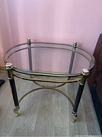 Front view of vintage oblong glass table showing clear glass top and metal frame with black legs and gold accents, caster wheels visible.