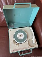 Green portable record player with lid open, showing turntable, tonearm, speaker grill, and controls on a maroon leather surface.