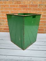 Green square heavy metal bin with rust and wear, shown on a gray wooden deck against a brick wall background.