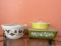 View showing three dishes: white patterned mixing bowl, green floral casserole with lid, and yellow round casserole with lid on top