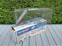 Metal wire cage trap for live animal capture positioned next to its original product packaging box on a wooden surface with greenery background.