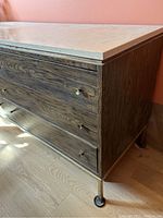 Side angle view of dresser showing faux wood drawer fronts, chrome legs with wheels, and marble-like surface top.