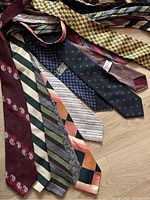 Top-down photo showing a spread of 9 vintage colorful ties laid out on a wood floor. Colors include burgundy with paisley, green and cream stripes, yellow checkered, and other patterns.