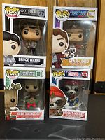 Four boxed Funko Pop figures arranged on a wooden surface: Bruce Wayne, Star-Lord, Holiday Dancing Groot, and Rocket (Holiday). Each in original packaging featuring character art and worn edges on boxes visible.