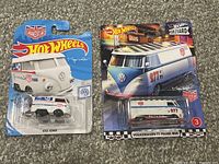 Photo shows two Hot Wheels Volkswagen T1 vans in original blister card packaging; Kool Kombi white van with Union Jack flag graphic and Boulevard Volkswagen T1 panel bus with blue and white checkerboard roof and '277 CI' markings.
