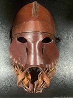 Front view of the brown leather mask showing eye holes and fringed bottom.